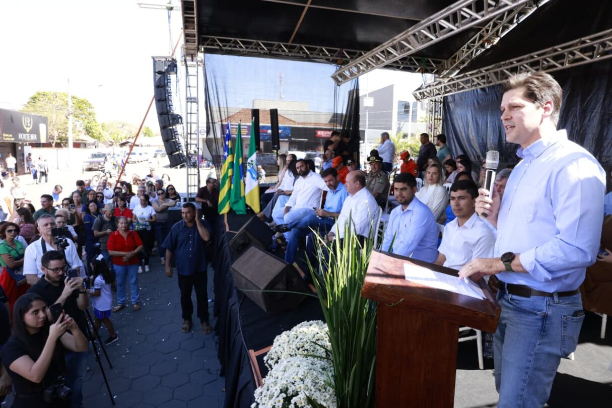 Imagem Daniel Vilela falando ao microfone, pessoas aparecem ao fundo em cima de palco