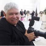 Imagem Fotógrafo Tião Marks, ele está sorrindo e segura uma maquina fotográfica