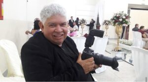 Imagem Fotógrafo Tião Marks, ele está sorrindo e segura uma maquina fotográfica