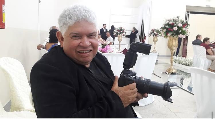 Imagem Fotógrafo Tião Marks, ele está sorrindo e segura uma maquina fotográfica