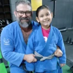 Imagem homem e criança de quimono sorrindo, ele está segurando a faixa do quimono da criança
