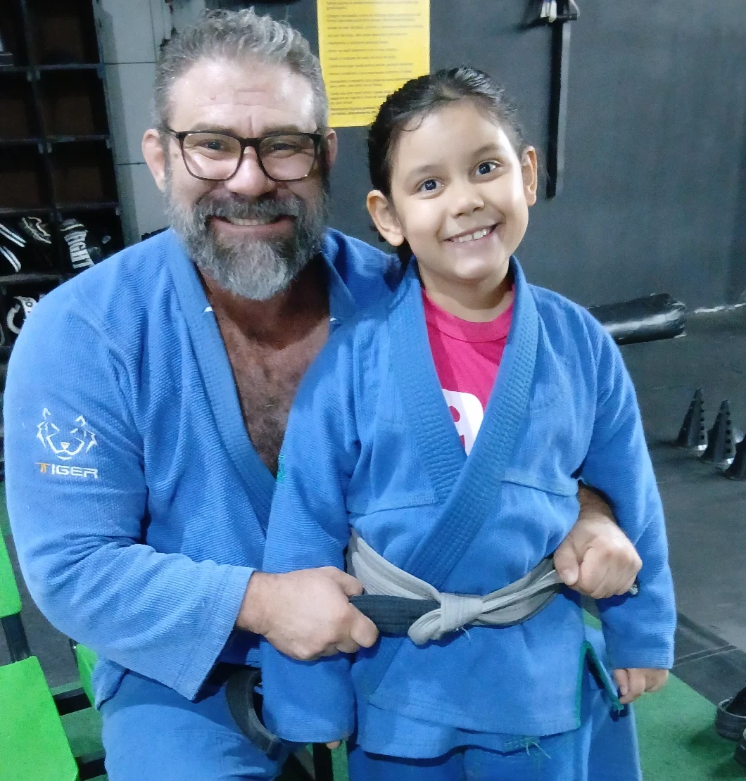 Imagem homem e criança de quimono sorrindo, ele está segurando a faixa do quimono da criança