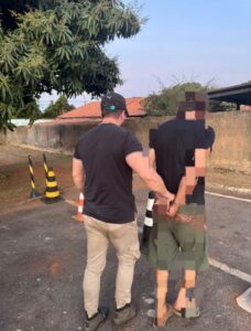 Imagem homem preso algemado com as mãos para traz, sendo conduzido por policial