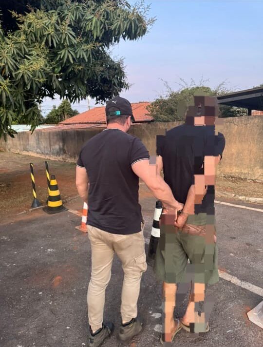 Imagem homem preso algemado com as mãos para traz, sendo conduzido por policial