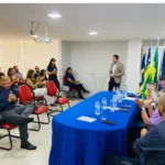 Imagem pessoas sentadas em cadeiras, a frente estão pessoas em uma mesa
