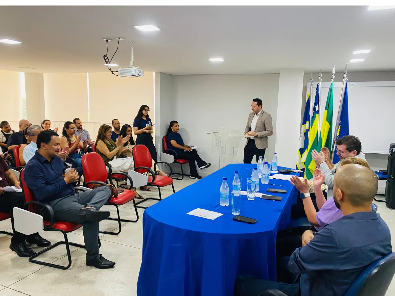 Imagem pessoas sentadas em cadeiras, a frente estão pessoas em uma mesa