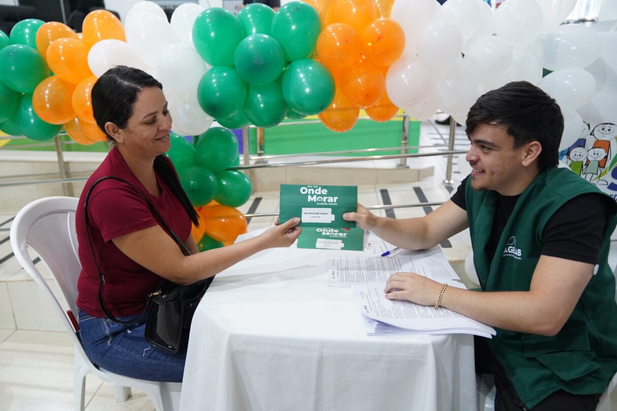 Imagem mulher e homem em uma mesa, ele está entregado um papel na mão dela