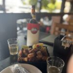 Imagem Cerveja e tira gosto no bar, uma garrafa de cerveja e dois copos pela metade no meio uma porção de torresmo com mandioca