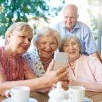 Imagem idosos sorrindo, três mulheres sentadas uma segurando o celular e um homem em pé