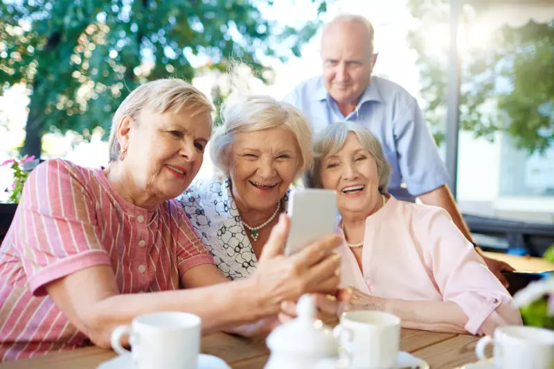 Imagem idosos sorrindo, três mulheres sentadas uma segurando o celular e um homem em pé