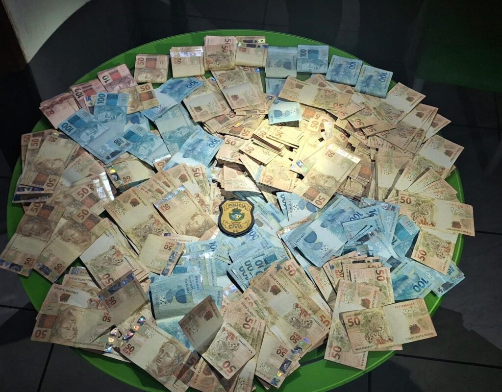 Imagem varias notas de cinquenta e cem reais em cima de uma mesa redonda, ao centro está o distintivo da Polícia Civil de Goiás