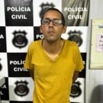 Imagem Jonathan Laurindo , ele está de camisa amarela com os braços para traz na delegacia de policia