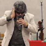 Imagem José Rico de Goiás. ele está de óculos escuro e terno branco, com a mão na orelha e segurando o microfone