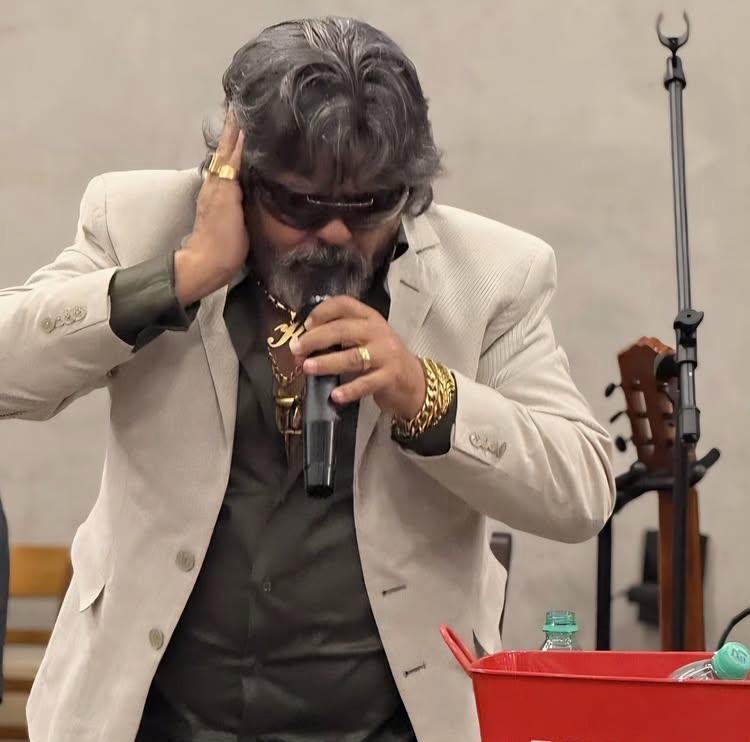 Imagem José Rico de Goiás. ele está de óculos escuro e terno branco, com a mão na orelha e segurando o microfone