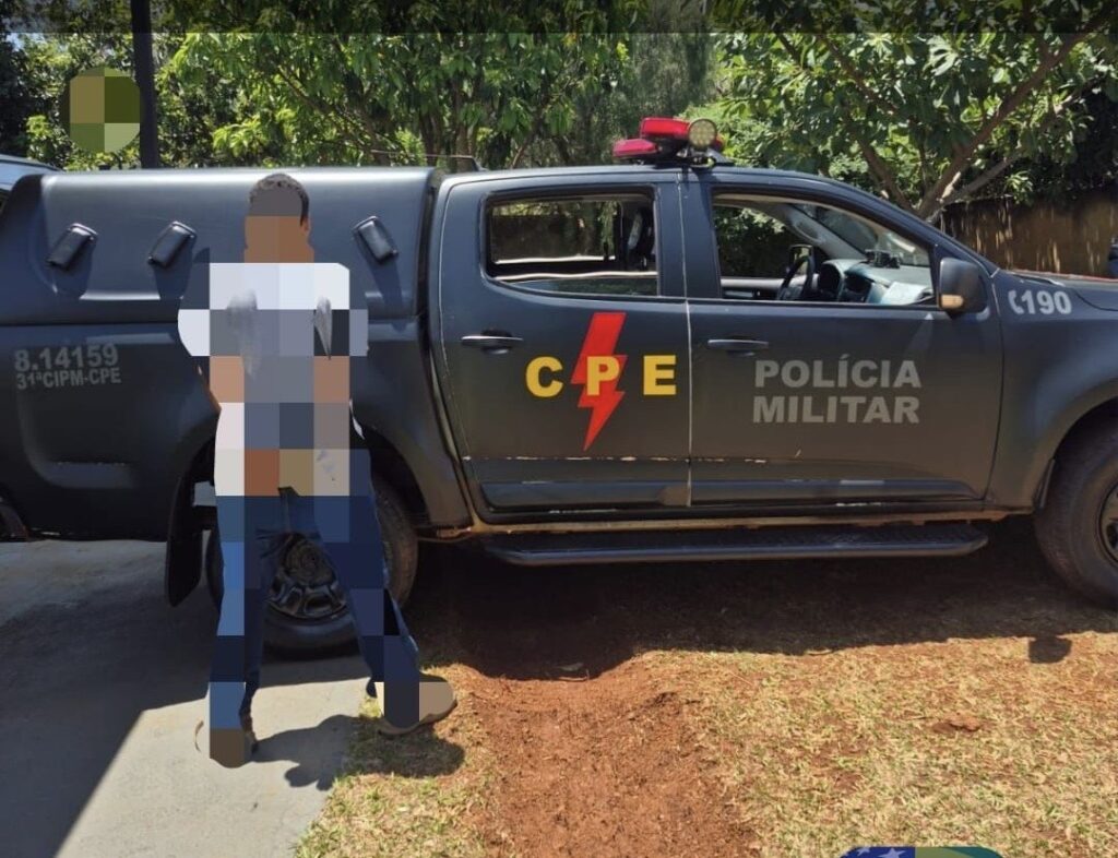 Imagem desfocada homem preso com as mãos para traz em frente a viatura da CPE