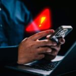 Imagem pessoas segurando um celular na frente de um computador e um sinal de alerta