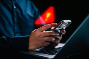 Imagem pessoas segurando um celular na frente de um computador e um sinal de alerta