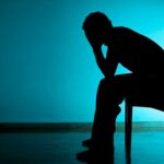 Imagem toda escura, tipo sombra, de uma pessoa sentada em uma cadeira com as duas mãos na cabeça