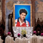 Imagem quadro com retrato de Carlo Acutis, ao redor estão padres e membros da Igreja Catolica