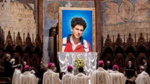 Imagem quadro com retrato de Carlo Acutis, ao redor estão padres e membros da Igreja Catolica