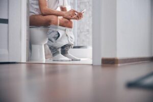 Imagem homem com as caças abaixadas sentado no vaso sanitário e mexendo no celular