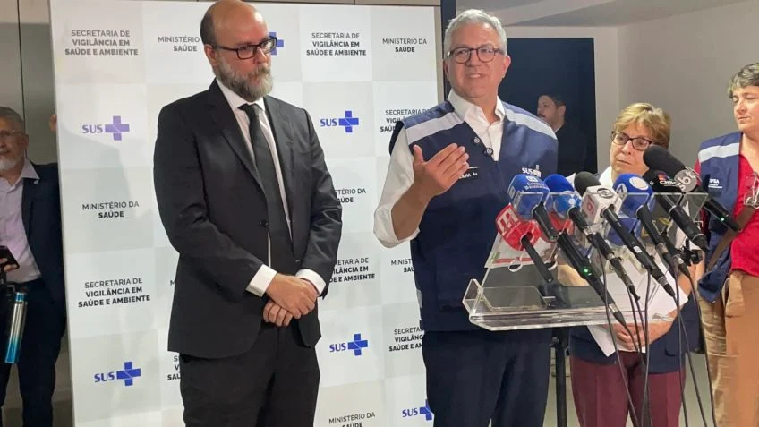 Imagem entrevista coletiva Ministro da Saúde, ele está de colete azul com vários microfones a frente