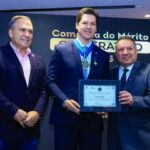Imagem Daniel Vilela segura uma placa de homenagem ladeado por duas pessoas