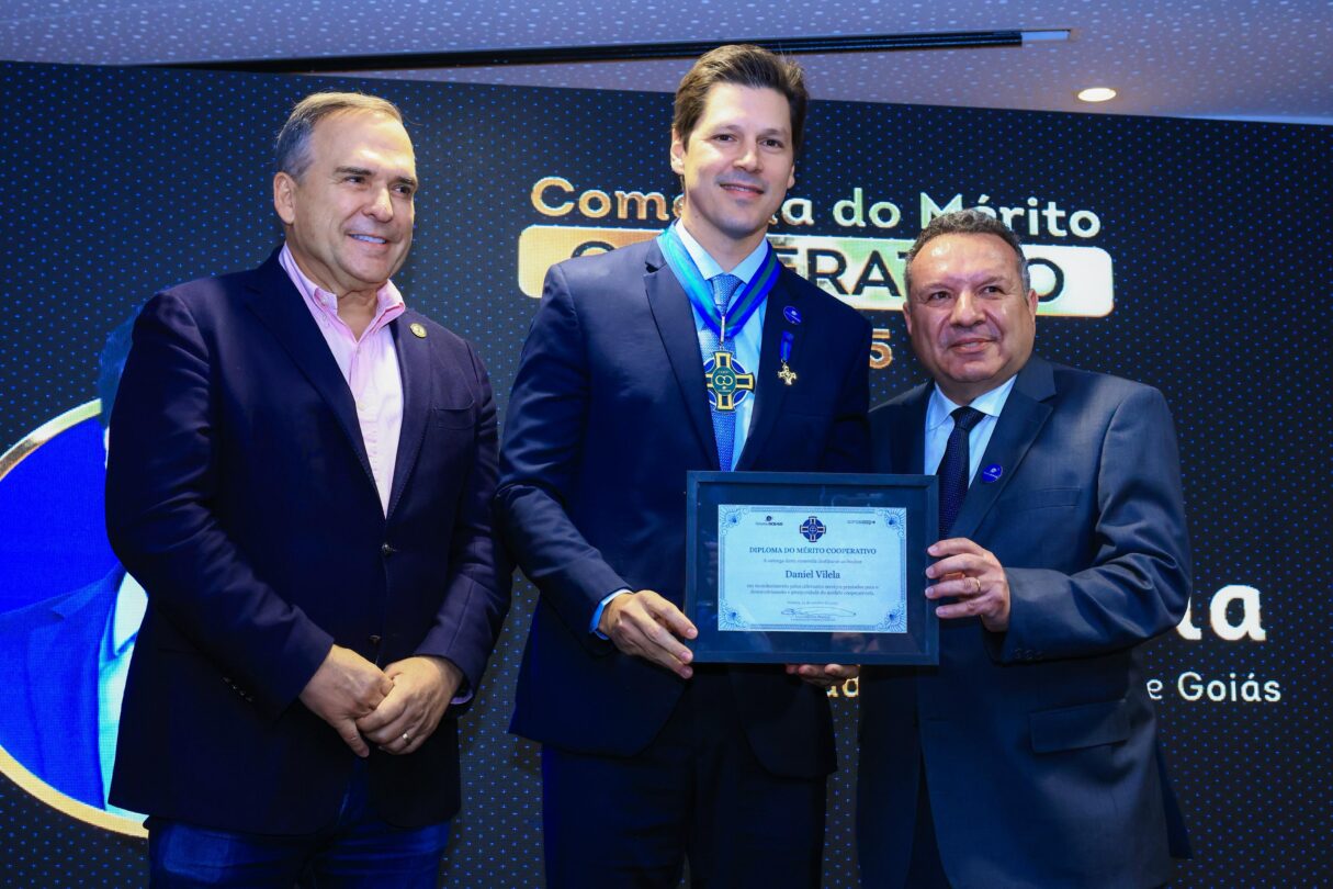 Imagem Daniel Vilela segura uma placa de homenagem ladeado por duas pessoas