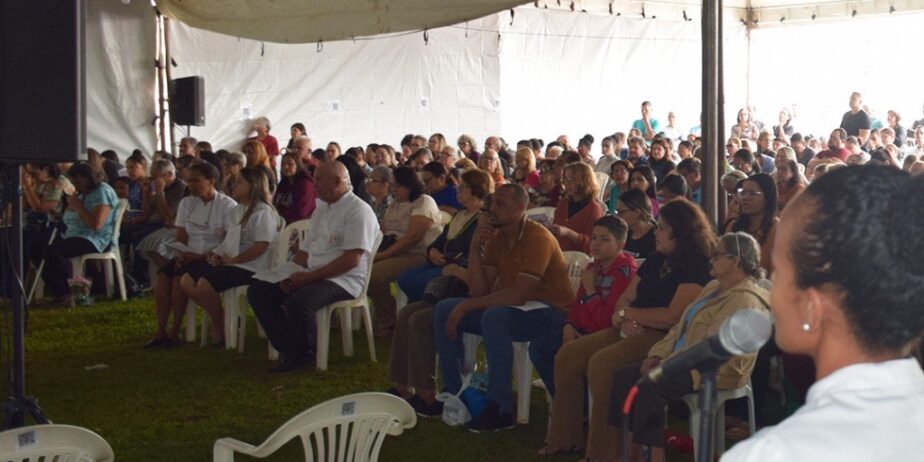 Imagem várias pessoas sentadas em baixo de uma atenda, a frente uma mulher com microfone