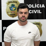 Imagem homem de camisa branca com os braços para traz preso na delegacia