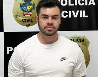 Imagem homem de camisa branca com os braços para traz preso na delegacia