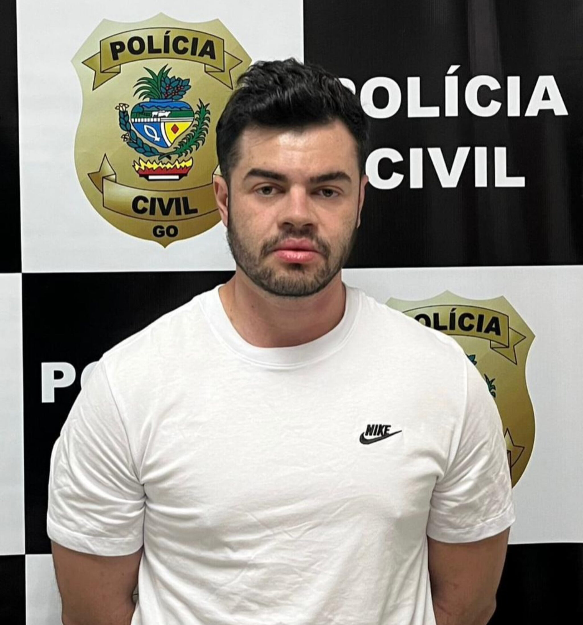 Imagem homem de camisa branca com os braços para traz preso na delegacia