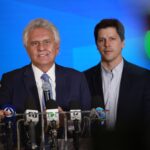 Imagem dois homens de terno e gravata um fala ao microfone