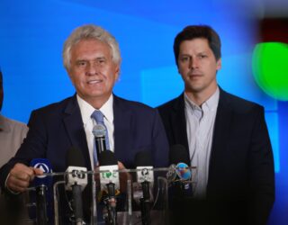 Imagem dois homens de terno e gravata um fala ao microfone