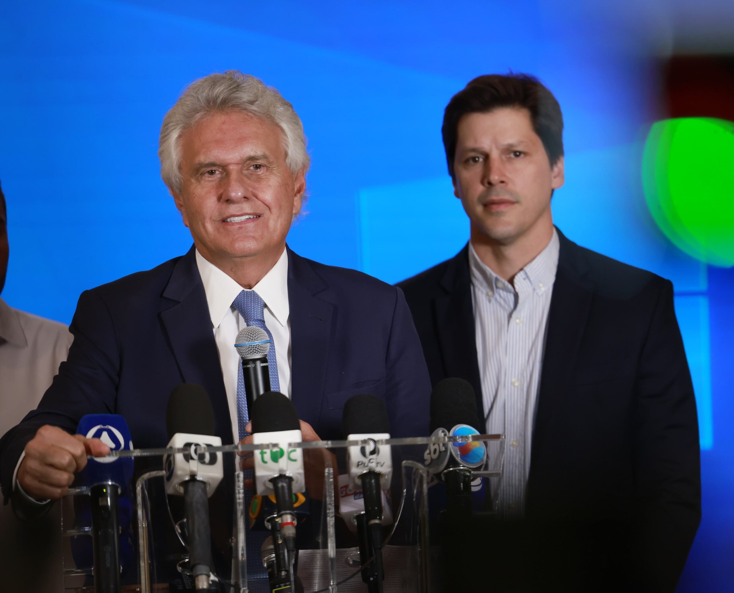 Imagem dois homens de terno e gravata um fala ao microfone