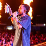 Imagem homem segurando microfone e um copo na mão, ao fundo varias pessoas