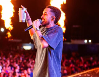 Imagem homem segurando microfone e um copo na mão, ao fundo varias pessoas