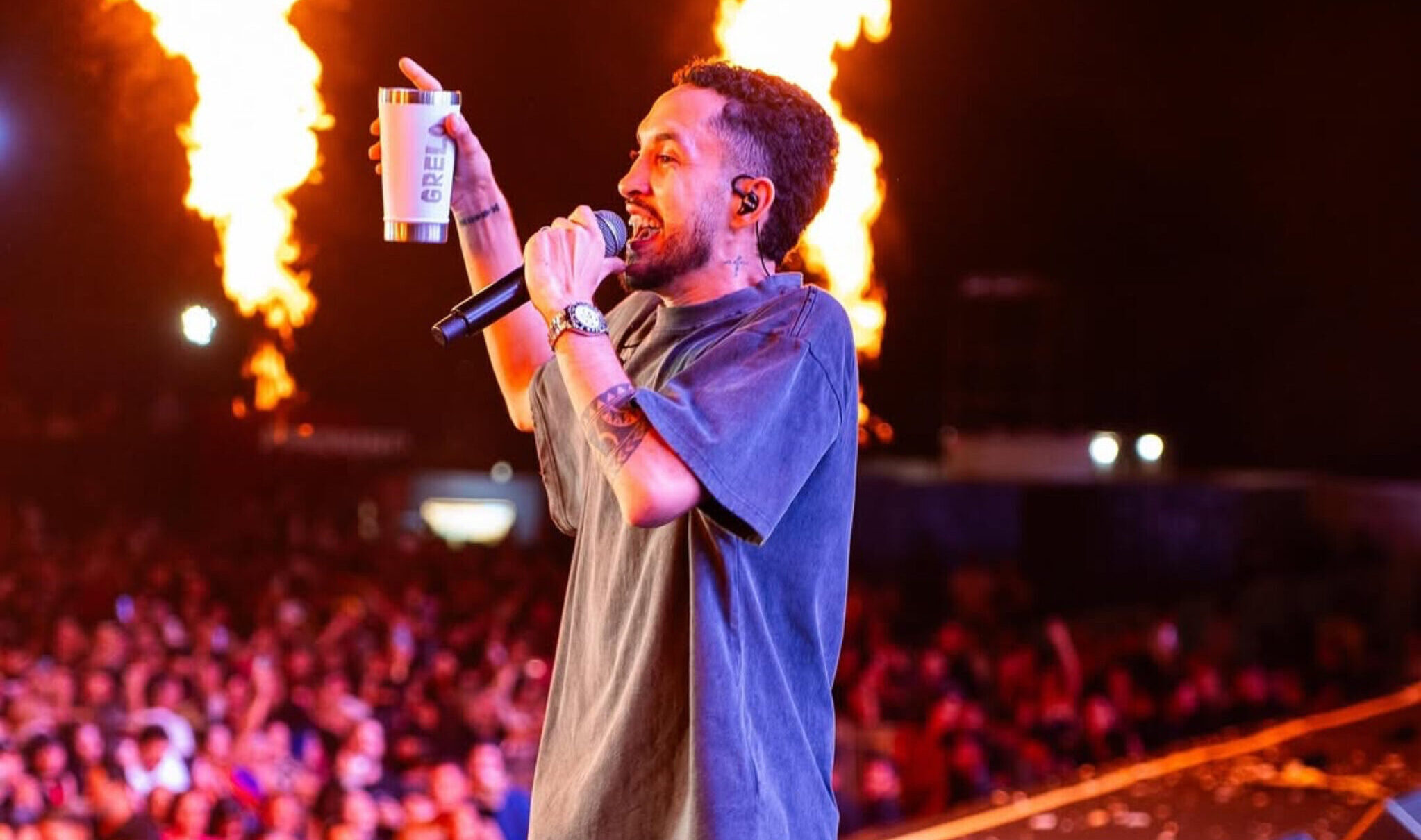 Imagem homem segurando microfone e um copo na mão, ao fundo varias pessoas