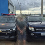 Imagem desfocada homem preso ao fundo camionete e viatura da policia