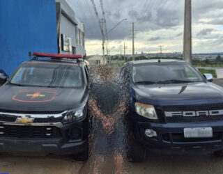 Imagem desfocada homem preso ao fundo camionete e viatura da policia