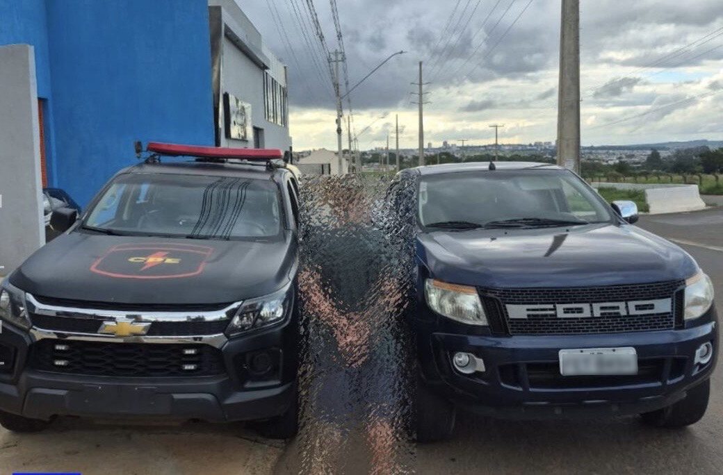 Imagem desfocada homem preso ao fundo camionete e viatura da policia