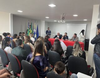 Imagem publico lota sala a frente três pessoas em uma mesa