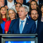 Imagem homem de terno e grava discursando ao fundo pessoas acompanham