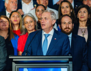 Imagem homem de terno e grava discursando ao fundo pessoas acompanham