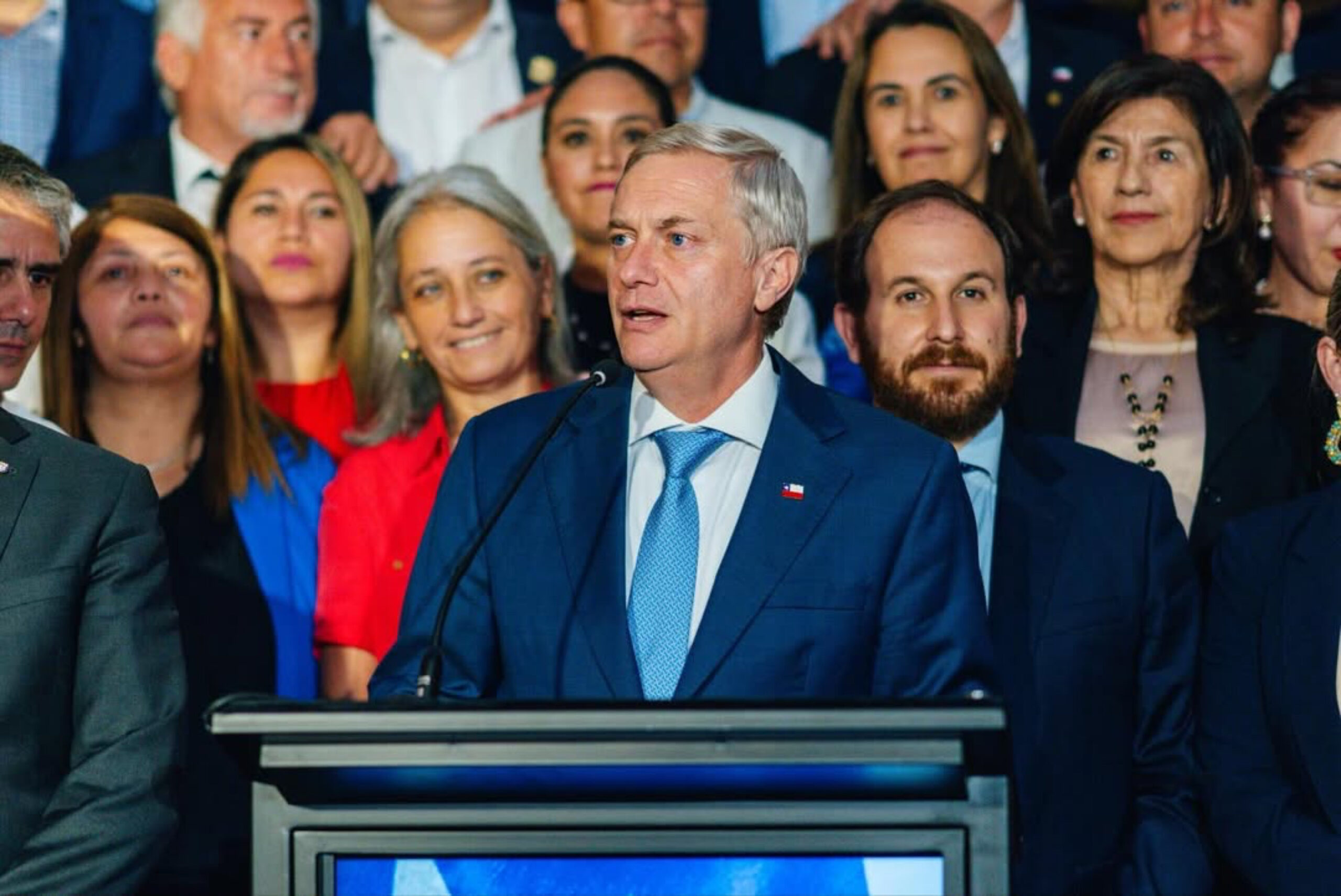 Imagem homem de terno e grava discursando ao fundo pessoas acompanham