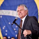 Imagem homem sorridente com microfone a frente ao fundo a bandeira do brasil