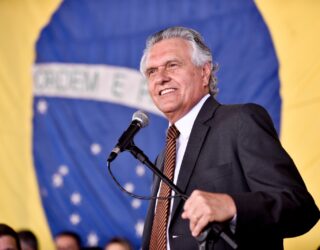 Imagem homem sorridente com microfone a frente ao fundo a bandeira do brasil