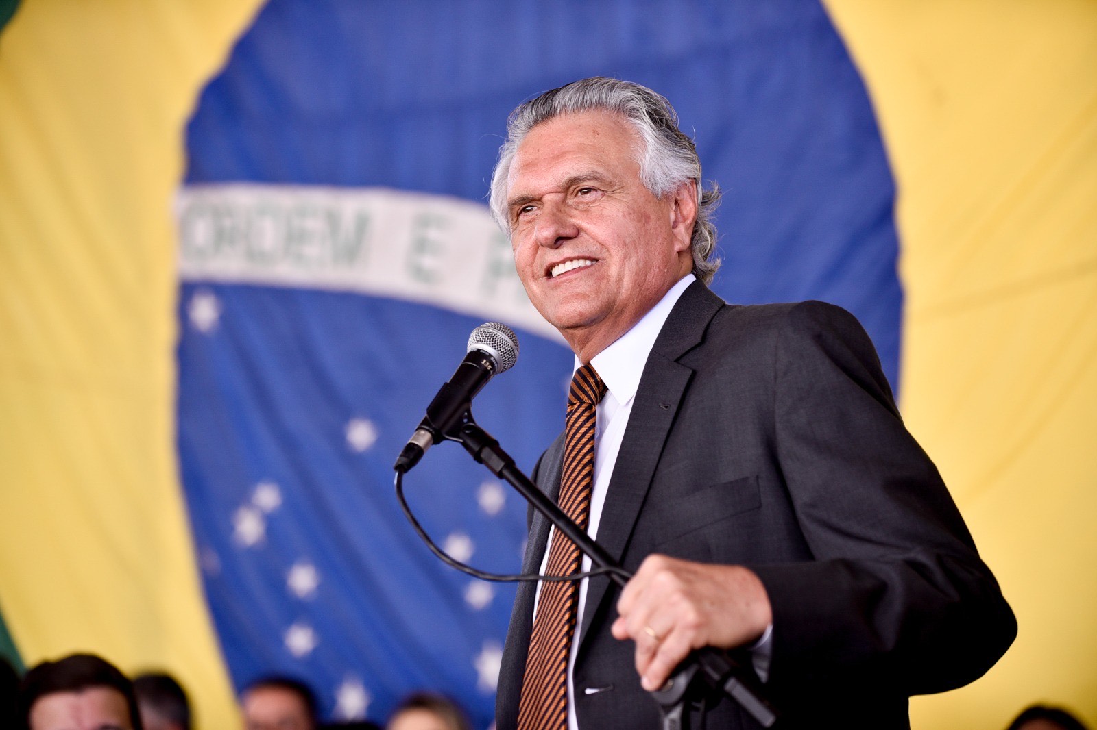 Imagem homem sorridente com microfone a frente ao fundo a bandeira do brasil