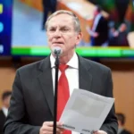 Imagem homem de terno e gravata vermelha segurando papel em frente microfone