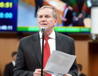 Imagem homem de terno e gravata vermelha segurando papel em frente microfone